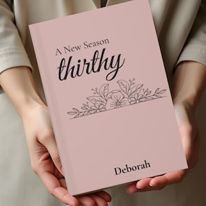 Może przedstawiać: Różowy notatnik z napisami "A New Season" i "thirthy" czarnym pismem. Notatnik ma kwiatowy wzór i imię "Deborah" na dole. Notatnik jest trzymany na tle beżowego tła.