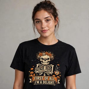 Może przedstawiać: Czarny t-shirt z grafiką szkieletu w wianku z kwiatów, ze skrzyżowanymi ramionami i parującą filiżanką kawy. Wzór zawiera małe dynie oraz napis "FIRST OF ALL, I'M A DELIGHT" wydrukowany na banerze u dołu koszulki.