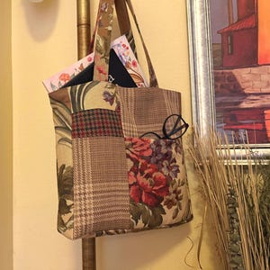 May include: A patchwork tote bag with various fabric patterns, including floral, plaid, and leaf designs. The bag has a brown leather strap and contains books and a pair of black glasses. The bag is hanging on a gold pole.