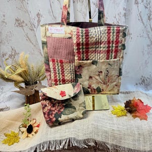 May include: A quilted tote bag featuring a patchwork design of floral, plaid, and textured fabric panels in shades of red, green, and cream, accompanied by a matching small flap pouch with a red flower-shaped button closure.