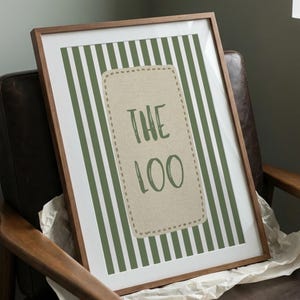 May include: A framed wall art print featuring a vertical green and white striped background. Centered is a beige rectangular patch with a stitched border containing the dark green text THE LOO in a hand-drawn style font within a wooden frame.