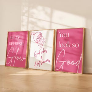 May include: Three framed wall art prints with gold frames. The prints feature pink backgrounds with white text. One says "Relax. Refresh. Glow", another has a shower illustration and says "Good vibes & Hot showers", and the last says "You look so Good."