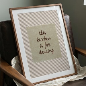 May include: A framed wall art piece featuring a beige and white vertical pinstripe background. Centered is a rectangular piece of textured tan fabric with frayed edges, displaying the dark brown text: this kitchen is for dancing.