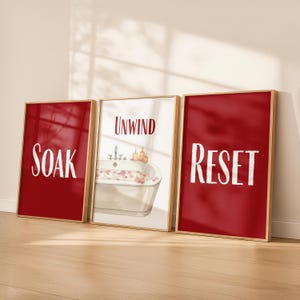 Könnte beinhalten: Ein Set aus drei gerahmten Wandbildern mit hellem Holzrahmen. Das linke Bild ist dunkelrot mit dem weißen Schriftzug SOAK. Das mittlere Bild ist weiß mit dem roten Schriftzug UNWIND und einer Badewanne. Das rechte Bild ist dunkelrot mit dem weißen Schriftzug RESET.