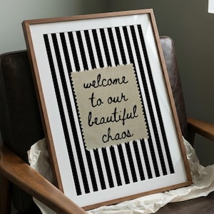 Könnte beinhalten: Ein gerahmtes Wandbild mit schwarz-weißem vertikalem Streifenhintergrund und einem mittigen beigen Stoffaufnäher. Der Aufnäher hat gezackte Ränder und zeigt den schwarzen handschriftlichen Text "welcome to our beautiful chaos" in Schreibschrift.