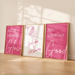 Puede incluir: Tres láminas decorativas rectangulares en marcos de madera clara. La lámina izquierda es rosa con texto blanco RELAX. REFRESH. Glow. La central es blanca con un icono de ducha rosa y el texto Good vibes & Hot showers. La derecha es rosa con texto blanco YOU LOOK SO Good.