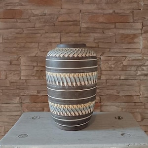 May include: A dark brown ceramic vase with horizontal white stripes and textured patterns. The vase has a cylindrical shape with a slightly wider base and a narrower neck. The textured sections feature a repeating leaf-like design in shades of blue and yellow.