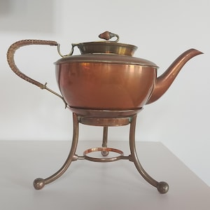 Könnte beinhalten: Kupfer-Teekanne mit geflochtenem Griff und Messingdeckel. Die Teekanne steht auf einem Messingständer mit gebogenen Beinen und einem kreisförmigen Sockel. Sie hat einen Ausguss und einen kleinen Knopf auf dem Deckel.