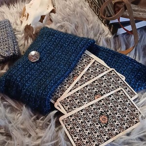 Puede incluir: Una funda de cartas de tarot de ganchillo azul oscuro con un botón plateado, que contiene una baraja de cartas de tarot. Las cartas tienen un diseño floral con una rosa en el centro. La escena incluye una calavera, libros y una vela.