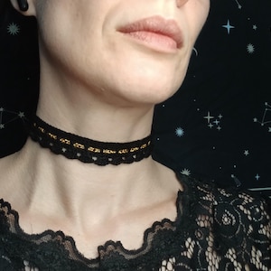 Può includere: Una collana girocollo all'uncinetto nera con un bordo smerlato e un dettaglio a catena dorata. La collana è indossata intorno al collo. La persona indossa un top di pizzo nero.