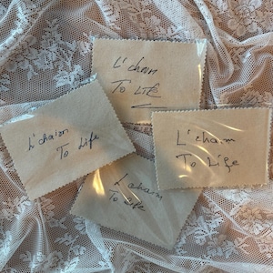 May include: Four rectangular beige fabric patches with pinked edges, each individually sealed in clear plastic. Each patch features the handwritten black ink text L'chaim To Life. The fabric has a visible woven texture and a natural light tan color.