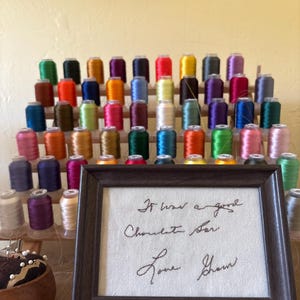 May include: A display of colorful thread spools in various shades, arranged on a wooden rack. A framed piece of art with handwritten text that reads "It was a good Chocolate Bar Love Shawn" is in the foreground. A pin cushion is also visible.