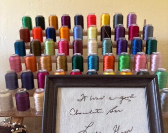 May include: A display of colorful thread spools in various shades, arranged on a wooden rack. A framed piece of art with handwritten text that reads "It was a good Chocolate Bar Love Shawn" is in the foreground. A pin cushion is also visible.