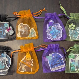 May include: Assortment of small, sheer drawstring bags in black, orange, purple, and green. Each bag features a themed sticker, including ghosts, haunted houses, and cartoon characters. The bags are tied with matching colored ribbons.