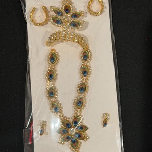 May include: A gold-coloured jewellery set comprising a necklace, earrings, and a headpiece. The necklace and headpiece are decorated with clear rhinestones and peacock feather designs in blue, green, and orange. The earrings are small, matching the necklace's design.