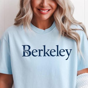 Puede incluir: Camiseta azul claro con la palabra "Berkeley" impresa en azul marino. La camiseta tiene mangas cortas y cuello redondo. Un suéter blanco está sobre el brazo. Un cactus está en el fondo.