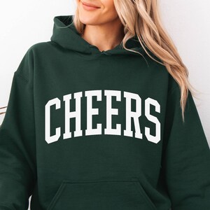 May include: Dark green hoodie with the word "CHEERS" in large, white, block letters across the chest. The hoodie has a front pocket and a hood. The person in the image is wearing light blue jeans.