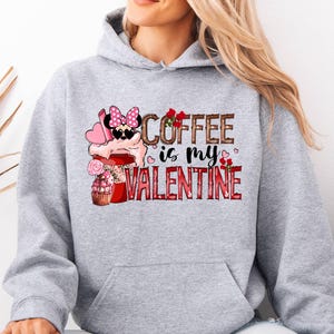 Peut inclure: Un sweat-shirt gris avec l'inscription "COFFEE is my VALENTINE" sur le devant. Le motif comprend une tasse de café avec un nœud, des cœurs et des roses. Le sweat-shirt a une poche avant et une capuche.