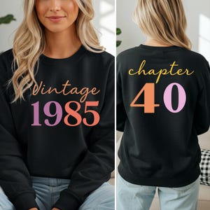 Può includere: Felpa nera con la scritta "Vintage 1985" in arancione e viola sul davanti. Il retro della felpa dice "chapter 40" in giallo, arancione e viola. La felpa è in stile casual.