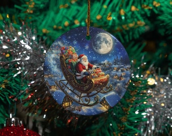Op de afbeelding: Ronde kerstversiering met de kerstman in zijn slee, vol cadeaus, vliegend over een besneeuwd dorp onder een volle maan. De ornament heeft een gouden touwtje om op te hangen.