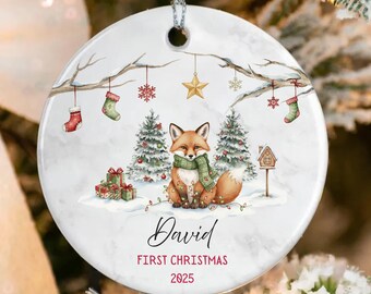 May include: A circular Christmas ornament with a watercolour design. It depicts a fox in a green scarf, Christmas trees, and the text "David FIRST CHRISTMAS 2025". The ornament is white with a silver hanging loop.