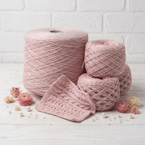 May include: A collection of pink yarn skeins and a knitted piece. The yarn is in various sizes, including a large spool and several smaller balls. The knitted piece has a cable pattern. Dried flowers and pearls are scattered around the yarn.