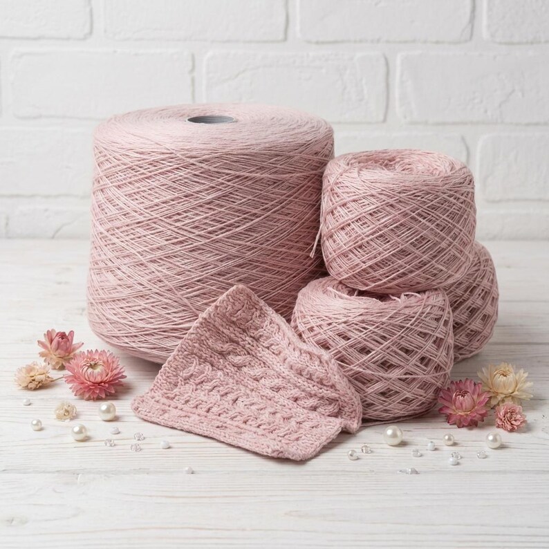 May include: A collection of pink yarn skeins and a knitted piece. The yarn is in various sizes, including a large spool and several smaller balls. The knitted piece has a cable pattern. Dried flowers and pearls are scattered around the yarn.