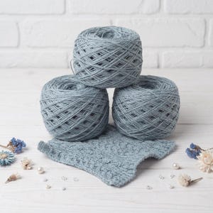 May include: Three balls of light blue yarn are stacked, with a knitted swatch in front. The yarn is a soft, muted blue color. The swatch has a textured pattern. The background is a white surface with decorative elements.