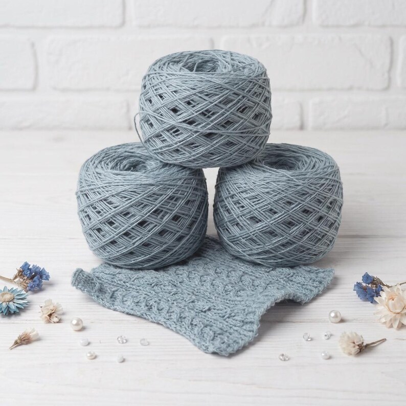 May include: Three balls of light blue yarn are stacked, with a knitted swatch in front. The yarn is a soft, muted blue color. The swatch has a textured pattern. The background is a white surface with decorative elements.