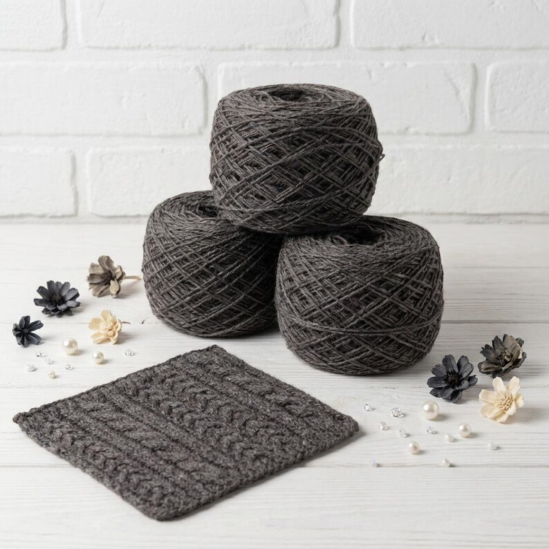 May include: Three gray yarn skeins stacked, with a knitted square in front. The yarn is a dark gray color, and the knitted square has a cable knit design. Decorative flowers and pearls are scattered around the yarn and knitted square.