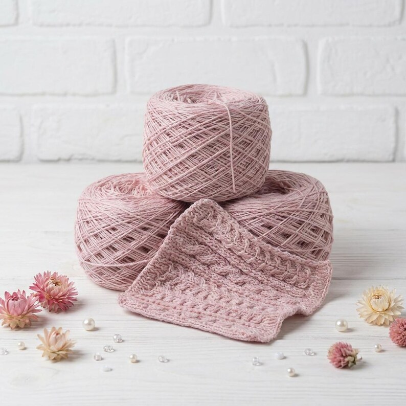 May include: Three balls of dusty rose yarn are stacked with a knitted swatch in the foreground. The yarn is tightly wound, and the swatch displays a textured cable knit pattern. Dried flowers and pearls are scattered around the yarn on a white surface.