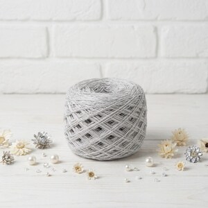 May include: A large ball of gray yarn sits on a white wooden surface, surrounded by decorative elements. The yarn is tightly wound, with a textured appearance. Scattered around are small pearls, clear crystals, and dried flowers, creating a delicate and artistic composition.