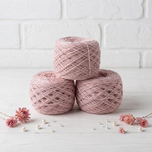 May include: Three balls of light pink yarn are stacked on a white surface. The yarn is arranged in a pyramid shape. Small pink flowers and pearls are scattered around the yarn balls. The background is a white brick wall.