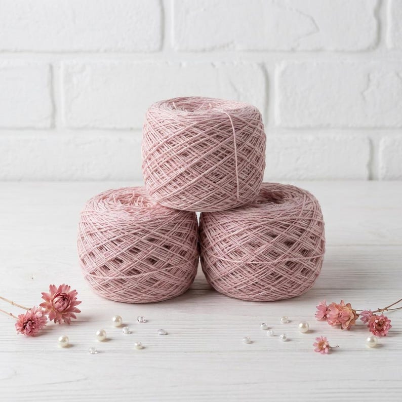 May include: Three balls of light pink yarn are stacked on a white surface. The yarn is arranged in a pyramid shape. Small pink flowers and pearls are scattered around the yarn balls. The background is a white brick wall.