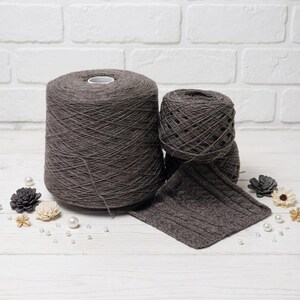May include: A large spool of dark gray yarn sits next to two smaller balls of yarn and a knitted swatch. The yarn is arranged on a white surface with decorative flowers and pearls.
