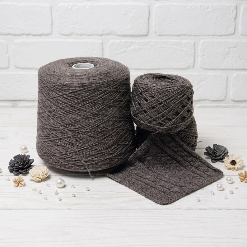 May include: A large spool of dark gray yarn sits next to two smaller balls of yarn and a knitted swatch. The yarn is arranged on a white surface with decorative flowers and pearls.