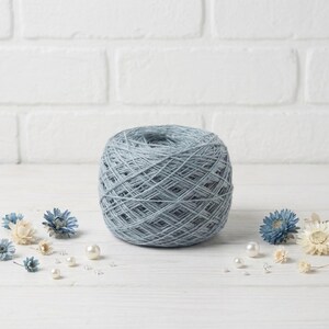 May include: A ball of light blue yarn sits on a white wooden surface, surrounded by small blue and white flowers and pearls. The yarn is tightly wound, showing the texture of the fibers. The background is a white brick wall.