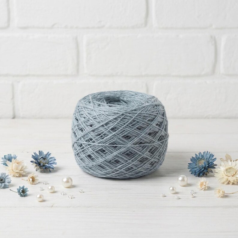 May include: A ball of light blue yarn sits on a white wooden surface, surrounded by small blue and white flowers and pearls. The yarn is tightly wound, showing the texture of the fibers. The background is a white brick wall.
