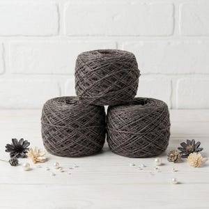 May include: Three dark gray yarn skeins stacked on a white wooden surface. The yarn is tightly wound, and the texture is visible. Decorative flowers and pearls are scattered around the base of the yarn. The background is a white brick wall.