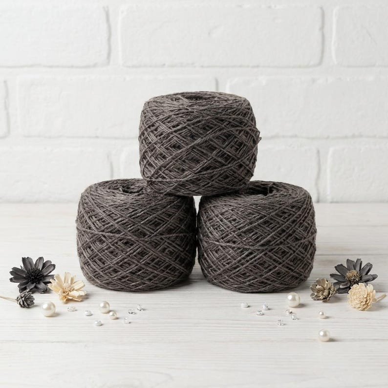 May include: Three dark gray yarn skeins stacked on a white wooden surface. The yarn is tightly wound, and the texture is visible. Decorative flowers and pearls are scattered around the base of the yarn. The background is a white brick wall.