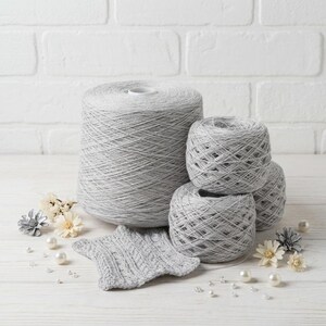 May include: A collection of gray yarn in various forms, including a large spool and several smaller balls. A small, knitted gray star-shaped swatch is also present, along with decorative pearls and flowers, suggesting a crafting theme.