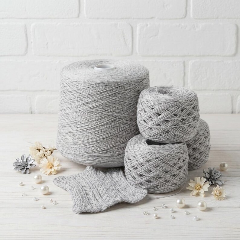 May include: A collection of gray yarn in various forms, including a large spool and several smaller balls. A small, knitted gray star-shaped swatch is also present, along with decorative pearls and flowers, suggesting a crafting theme.