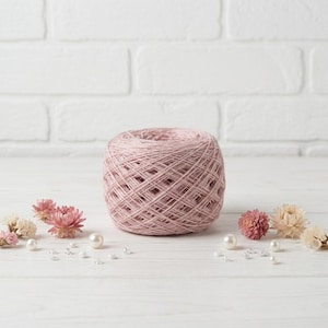 May include: A ball of pink yarn sits on a white wooden surface, surrounded by small pink and white flowers, pearls, and decorative beads. The yarn is a light pink color and appears to be made of a soft, natural fiber. The background is a white brick wall.