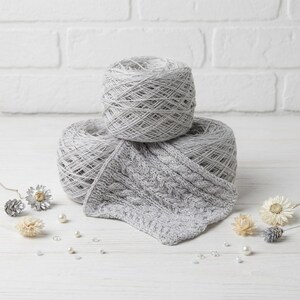 May include: Two gray yarn skeins with a knitted swatch. The yarn is a light gray color, and the knitted swatch has a cable knit pattern. The yarn and swatch are on a white wooden surface with decorative elements.