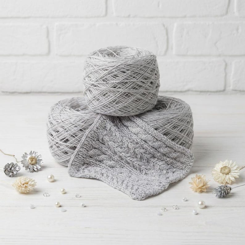 May include: Two gray yarn skeins with a knitted swatch. The yarn is a light gray color, and the knitted swatch has a cable knit pattern. The yarn and swatch are on a white wooden surface with decorative elements.