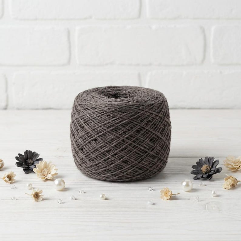 May include: A large, dark gray yarn ball sits on a white wooden surface, surrounded by decorative elements. Small, cream-colored and dark gray flowers, along with pearls and clear crystals, are scattered around the yarn, creating a craft-themed composition.