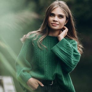 Könnte beinhalten: Ein grüner Zopfstrickpullover mit Rundhalsausschnitt und langen Ärmeln. Der Pullover wird mit einer dunklen Jeans und einem schwarzen Gürtel getragen. Der Hintergrund ist eine verschwommene Szene im Freien mit grünem Laub.