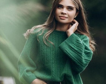 May include: A person wearing a green cable-knit sweater with long sleeves and a crew neck. The sweater is paired with dark denim jeans and a black belt. The background is a blurred outdoor scene with green foliage.
