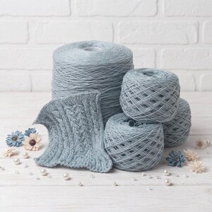 May include: A collection of light blue yarn skeins and a knitted swatch. The yarn is arranged with decorative flowers and pearls on a white wooden surface. The knitted swatch displays a cable knit pattern.