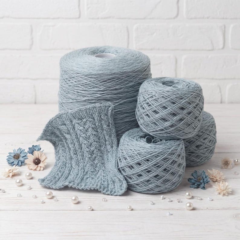May include: A collection of light blue yarn skeins and a knitted swatch. The yarn is arranged with decorative flowers and pearls on a white wooden surface. The knitted swatch displays a cable knit pattern.